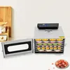 A stainless steel food dehydrator with multiple trays filled with sliced fruits, placed on a countertop next to a wooden cutting board and a jar of sugar cubes. A small bowl of dried fruit is in the foreground. Podgląd zdjęcia produktu