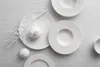 A collection of white Villeroy & Boch Manufacture Rock Blanc dinner plates and bowls arranged on a light gray textured surface, with decorative white branches and pomegranates. Produktbild-Vorschau
