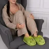 A person sitting in a chair wearing loungewear and Grinch-themed slippers. Produktbild-Vorschau