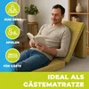 A man is reading a book while sitting on a folded mattress in a living room. Produktbild-Vorschau