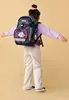 A young girl wearing a backpack stands with her arms outstretched against a plain background. Náhled obrázku produktu