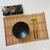 A bamboo placemat with a black bowl and chopsticks, along with small dishes containing spices and a sachet, arranged on a white surface. Produktbild-Vorschau