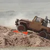 A brown off-road vehicle is driving over orange traction boards on a dirt hill, kicking up dust. Náhled obrázku produktu