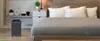 A bedroom scene with an air purifier next to a bed. Produktbild-Vorschau