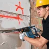 A man in a yellow hard hat and safety glasses is using a Bosch GBH 18V-26 Akku-Bohrhammer to break through a concrete wall. Red spray paint marks are visible on the wall. Produktbild-Vorschau