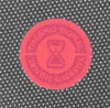 A close-up of a red circular stamp with an hourglass symbol in the center, set against a black background with white polka dots. Anteprima dell’immagine prodotto