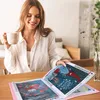 A woman is sitting at a table, holding a mug and looking at an open album with diamond paintings. Produktbild-Vorschau