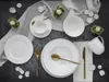 A flat lay of white porcelain dinnerware set on a dark textured surface with a sheer white cloth draped. Produktbild-Vorschau