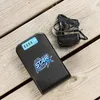 A black power bank with a blue logo and charging indicator lights sits on a wooden surface next to a black power adapter and cord. Produktbild-Vorschau