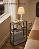A narrow side table with a lamp, cup, and phone on it, placed next to a bed in a room. Produktbild-Vorschau