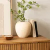 A beige ceramic vase with green branches sits on a wooden dresser next to two books. A small wooden candle is in the foreground. Náhled obrázku produktu