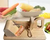 A wooden sausage slicer with a sausage being cut, with a blurred background of vegetables. Náhled obrázku produktu
