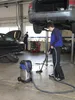 A man is vacuuming a wet floor in a car repair shop, with cars and tools in the background. Anteprima dell’immagine prodotto