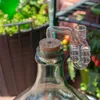 A close-up of a glass carboy with a cork and a glass fermentation lock, set against a blurred background of plants and tomatoes. Produktbild-Vorschau