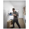 A man is using a cordless hammer drill to drill into a white wall in a room with wooden floors and kitchen cabinets in the background. Produktbild-Vorschau
