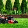 A lawnmower is shown on a freshly cut green lawn, with a rustic woven fence and evergreen trees in the background. Produktbild-Vorschau