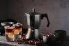 A still life arrangement of a Moka pot, two glasses of coffee, cherries, and coffee beans on a wooden surface. Náhľad obrázku produktu