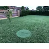A green manhole cover sits on a lush green lawn in a backyard, with a wooden fence and houses in the background. Produktbild-Vorschau