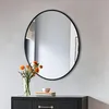 A round mirror with a black frame hangs on a light gray wall above a black dresser. A vase of orange flowers sits on the dresser to the left of the mirror. Podgląd zdjęcia produktu