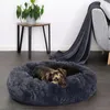 A dog is resting comfortably in a plush donut-shaped dog bed on a dark floor, with a side table and a blanket draped over a chair in the background. Anteprima dell’immagine prodotto