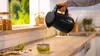 A person is pouring hot water from a black electric kettle into a glass cup with a tea infuser. Podgląd zdjęcia produktu