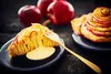 A slice of apple pie with custard and a whole apple pie are presented on dark plates, with whole apples in the background. Produktbild-Vorschau