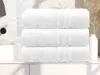 Three white towels are neatly stacked on a shelf next to a sink and some decorative reeds in a vase. Anteprima dell’immagine prodotto