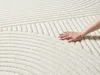 A hand is shown touching a textured rug with a geometric pattern. Produktbild-Vorschau