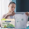 A woman is looking at a stained white t-shirt, holding a box of Heitmann Gallseife (gall soap) in the foreground. Produktbild-Vorschau