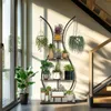 A multi-tiered plant stand filled with various potted plants, placed indoors near a large window and a staircase. Náhled obrázku produktu