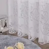 A close-up view of a white floral patterned curtain, with a fluffy gray rug and some decorative flowers in the foreground. Produktbild-Vorschau