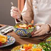 A person is eating a salad from a colorful, patterned bowl on a wooden table. Náhled obrázku produktu