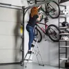 A woman is using a step ladder in a garage to reach bicycles mounted on the wall. Podgląd zdjęcia produktu
