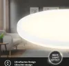 A close-up of a white LED panel with a backlight effect, set against a blurred background of a living room. Produktbild-Vorschau