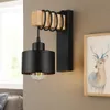 A modern wall lamp with a black shade and wooden accents is mounted on a wall next to a framed picture of a deer. Náhľad obrázku produktu