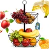 A two-tiered fruit basket filled with various fruits, set against a white background with some fruits appearing to float around it. Produktbild-Vorschau