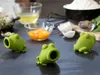Three green frog-shaped egg separators are arranged on a dark surface, with ingredients for baking in the background, including egg yolks, egg whites, flour, and herbs. Produktbild-Vorschau