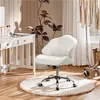 A white office chair with wheels is placed on a fluffy rug next to a white vanity table and a plant. Clothes are hanging on a rack in the background. Produktbild-Vorschau