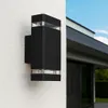 A black outdoor wall light mounted on a textured white wall, with a modern house and blue sky in the background. Náhľad obrázku produktu