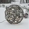 A black rattan sphere with embedded lights sits in the snow, surrounded by pine branches and snow-covered pinecones. Produktbild-Vorschau