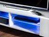 A close-up view of a white TV stand with blue LED lighting, showing a DVD player and a remote control. Produktbild-Vorschau