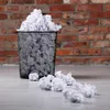 A black metal trash can filled with crumpled white paper, with more crumpled paper scattered on the floor in front of it, set against a brick wall. Náhled obrázku produktu
