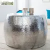 A silver hammered metal coffee table with decorative candle holders and a small glass cup on top, placed on a wooden floor in a bright room with a window. Produktbild-Vorschau