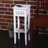 A white side table with a drawer and a shelf, placed next to a wooden chair, with a brick wall in the background and a plant on the left. Náhled obrázku produktu
