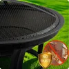 Close-up of a black fire pit with water droplets on its rim, set against a green grassy background. A circular inset shows a close-up of rusted metal with a red prohibition sign and a golden shield with text. Anteprima dell’immagine prodotto