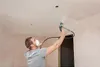 A man wearing a mask is spray painting a ceiling with a Wagner electric paint sprayer. Náhled obrázku produktu