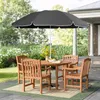 An outdoor patio set with a round table, four chairs, and a large dark gray umbrella, set on a rug with a backdrop of lush greenery and a house. Produktbild-Vorschau