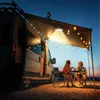 A couple sits at a table under an awning attached to a camper van, illuminated by string lights and a hanging lamp. Anteprima dell’immagine prodotto