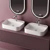 Two white ceramic sinks with silver faucets are shown on a pink vanity with a fluted texture. Two oval mirrors with integrated lighting are mounted on the pink wall behind the sinks. Voir les images du produit