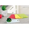 A set of four cutting boards in different colors (white, red, yellow, green) are arranged on a kitchen countertop. A green bell pepper sits on the white cutting board in the foreground, and two red tomatoes are visible to the left. A small potted plant is  Produktbild-Vorschau